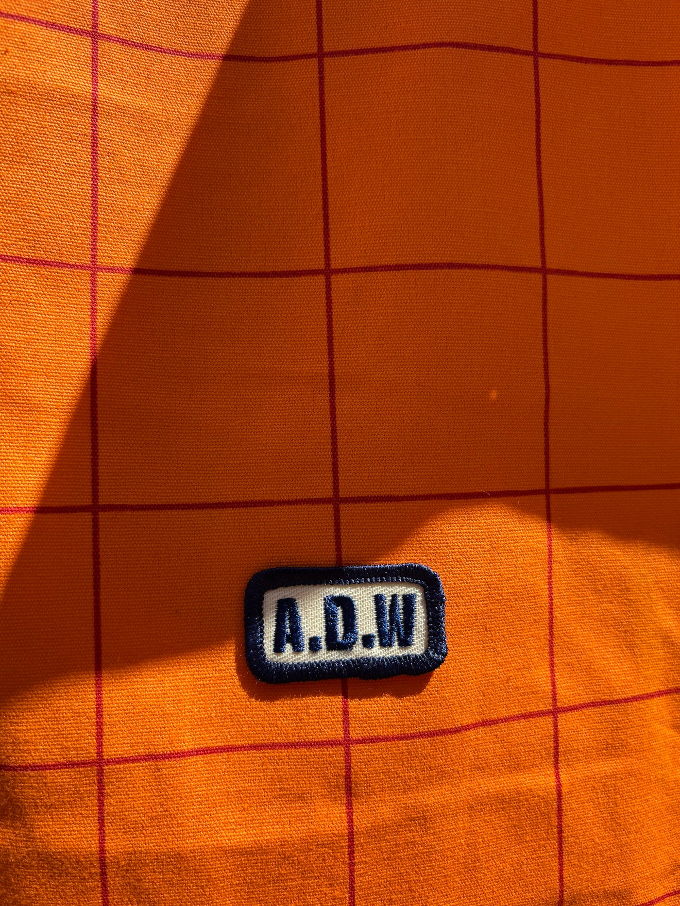 Fokus auf Baumwolltasche mit grafischem Grid-Print in Orange, gesticktem A.D.W. Patch und langen Henkeln – minimalistisches Streetwear-Design.